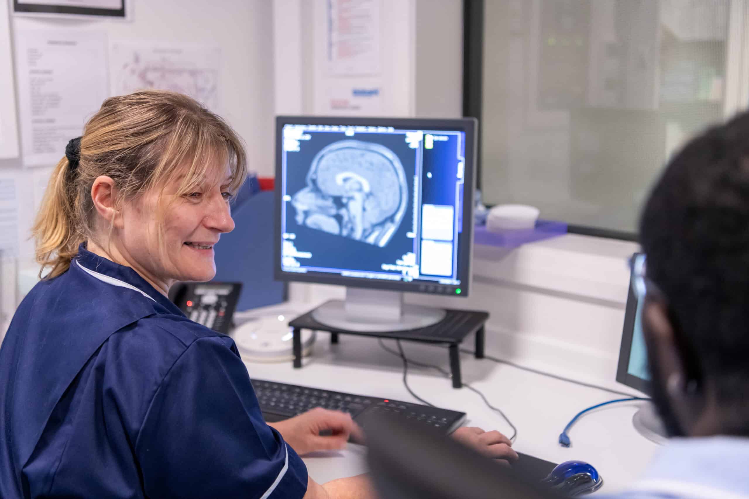 A nurse smiles and speaks to a colleague whilst a brain scan is shown on a computer screen in front of her