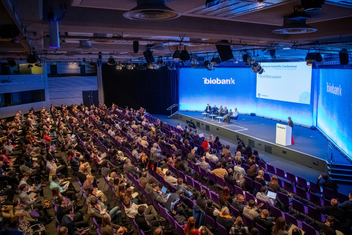 An overview shot of the UK Biobank scientific conference during a panel discussion with five people sitting on stage and rows of seats filled with attendees watching.