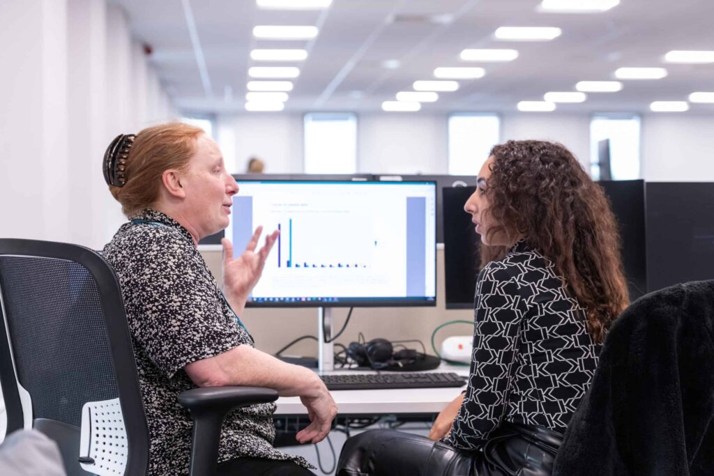 Two women sit talking in front of a computer screen displaying data