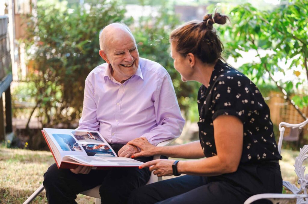An older man and younger woman sit looking at a photo album. Her hand is on his wrist supportively and he is smiling at her.