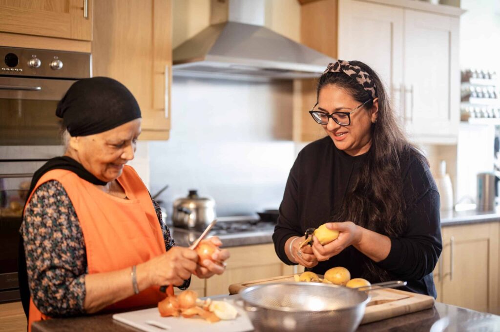 Two women in a kitchen chat and stand at a counter peeling vegetables together, smiling
