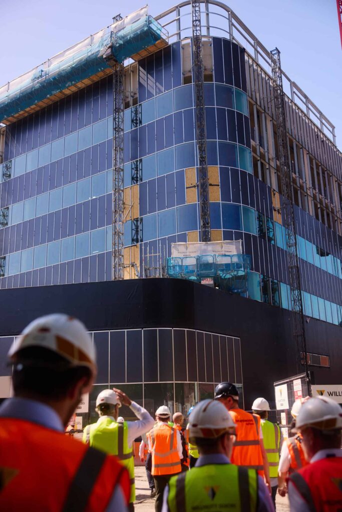 A crowd of people in hard hats and high visibility vests stand and look up at the top of a building being constructed