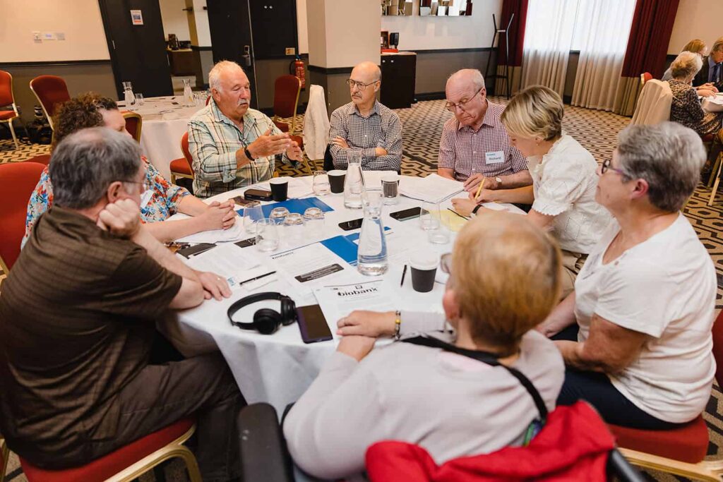 A group of UK Biobank participants sit round a table discussing something. They have UK Biobank branded materials in front of them.