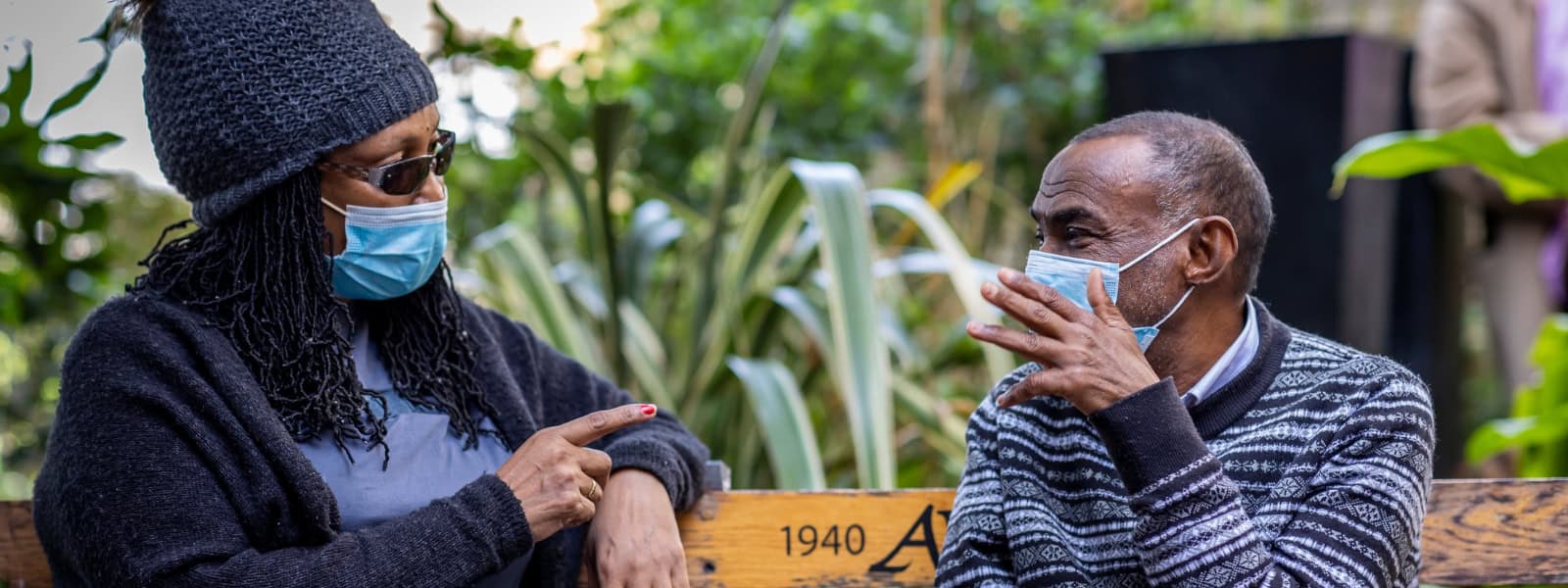 A man and a woman sit on a bench outside wearing masks