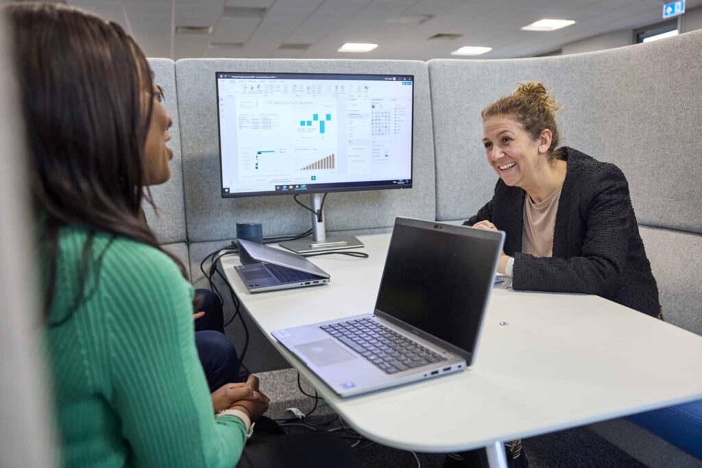 Two women sit and have a meeting in a booth, one has a laptop screen open and a monitor displays graphs and charts between them. They are smiling