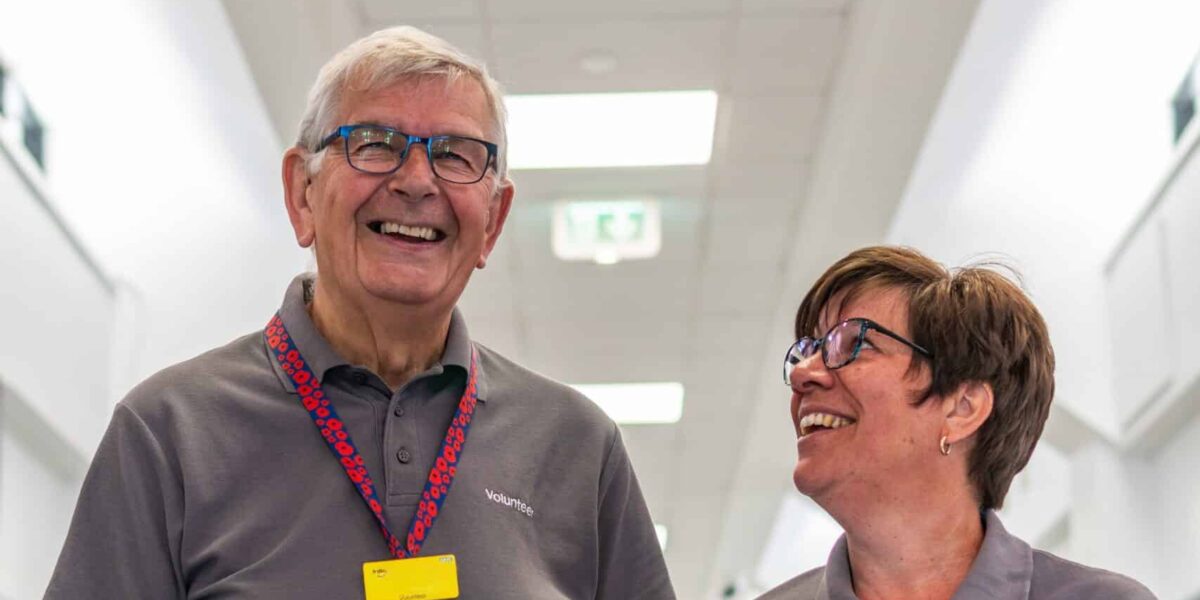 Two hospital volunteers smile whilst standing in a hospital corridor with the outpatients sign above their heads
