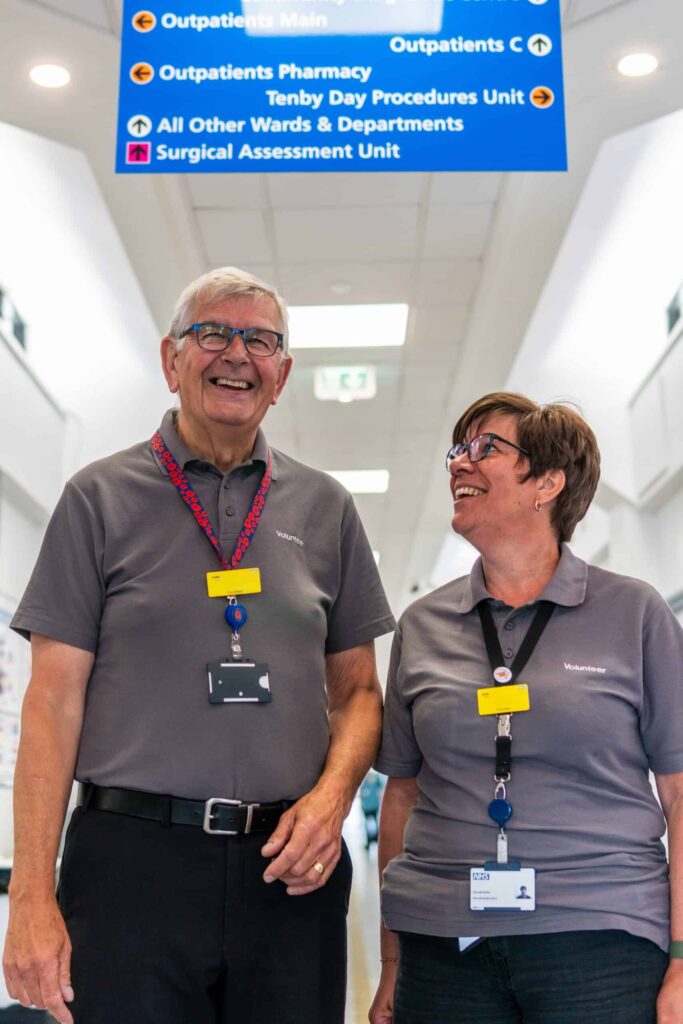 Two hospital volunteers smile whilst standing in a hospital corridor with the outpatients sign above their heads