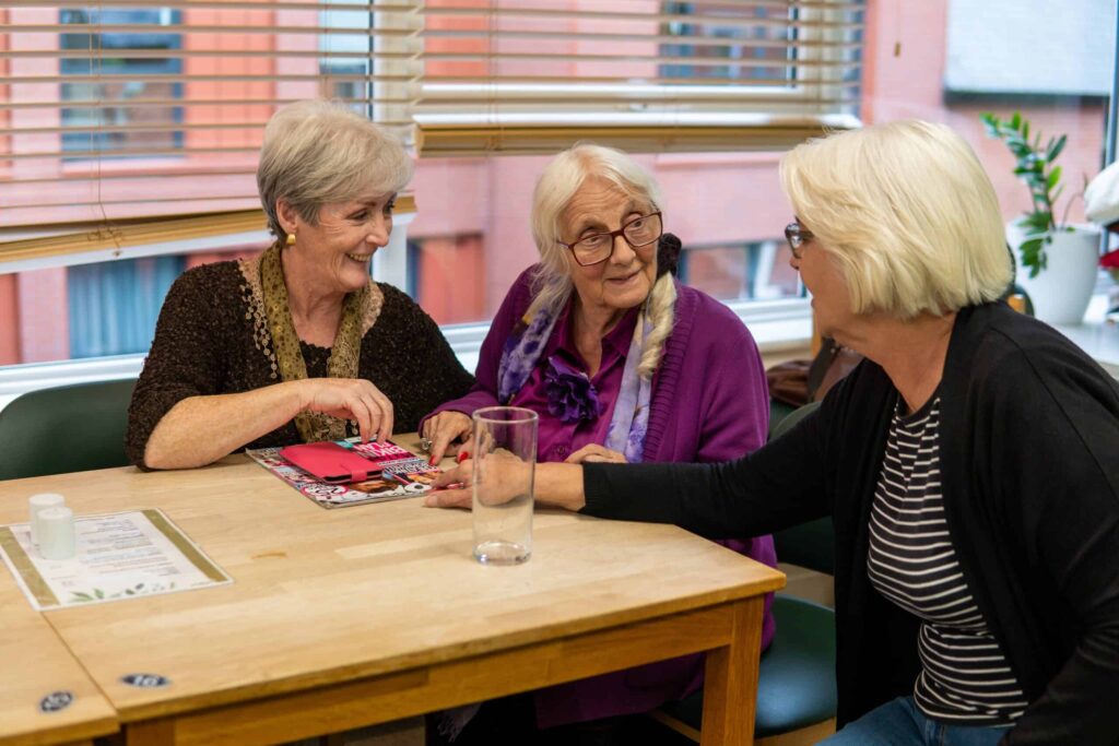 Three women sit and exchange conversation, smiling and pointing towards a magazine in front of them on the table
