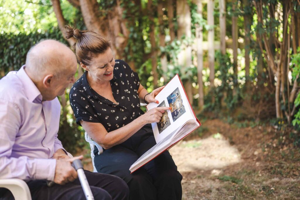 A woman sits with an older man outside, showing him old photos from a photo album.