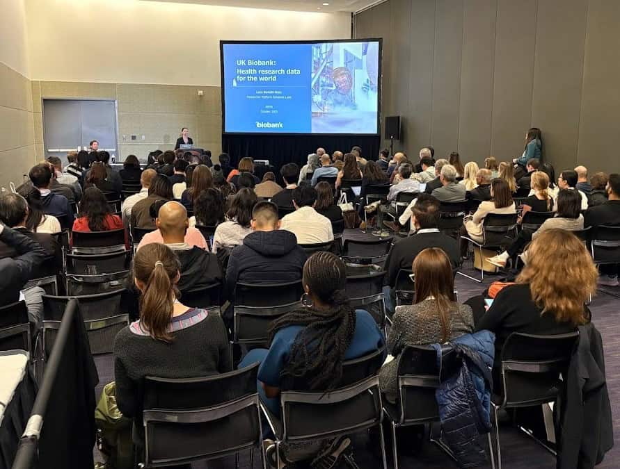 A UK Biobank staff member presents in front of a room full of people, sharing UK Biobank slides
