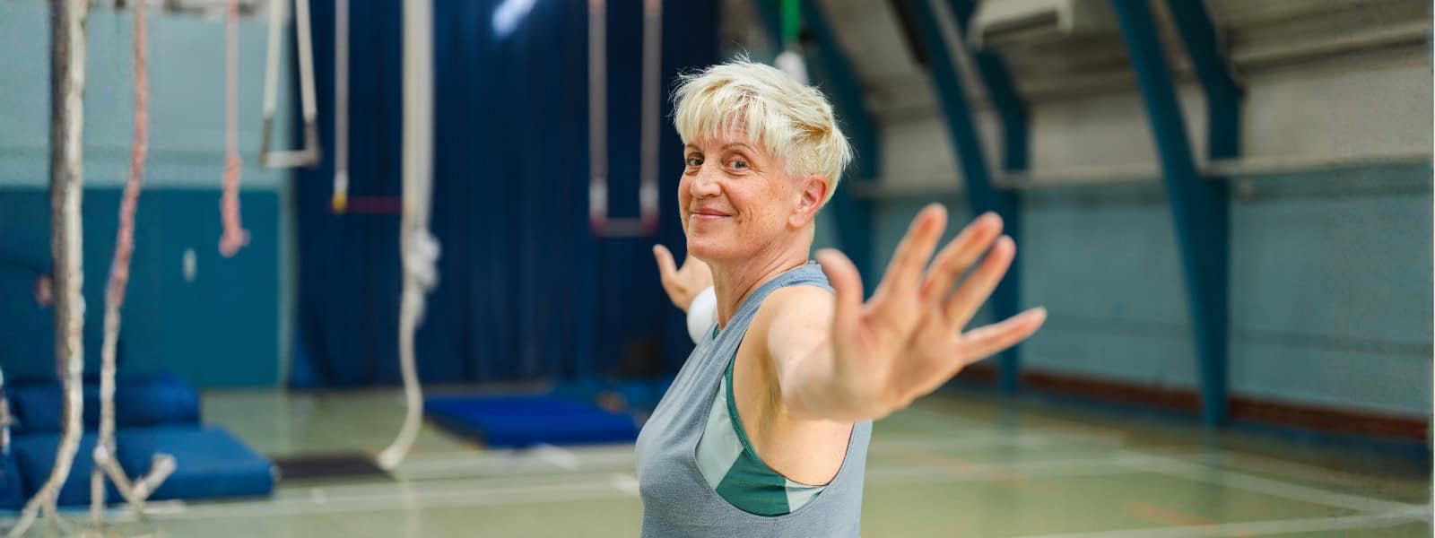 A woman in a sports hall wearing gym gear stretches her arms wide whilst smiling to the camera