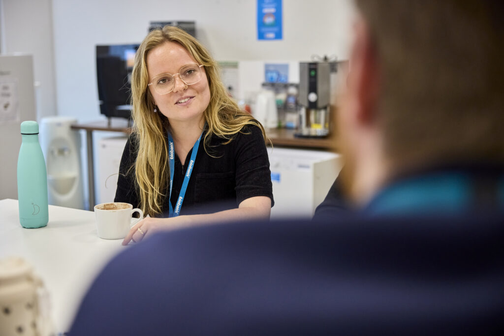 A woman sits talking to someone, with a cup of coffee and water bottle in front of them.
