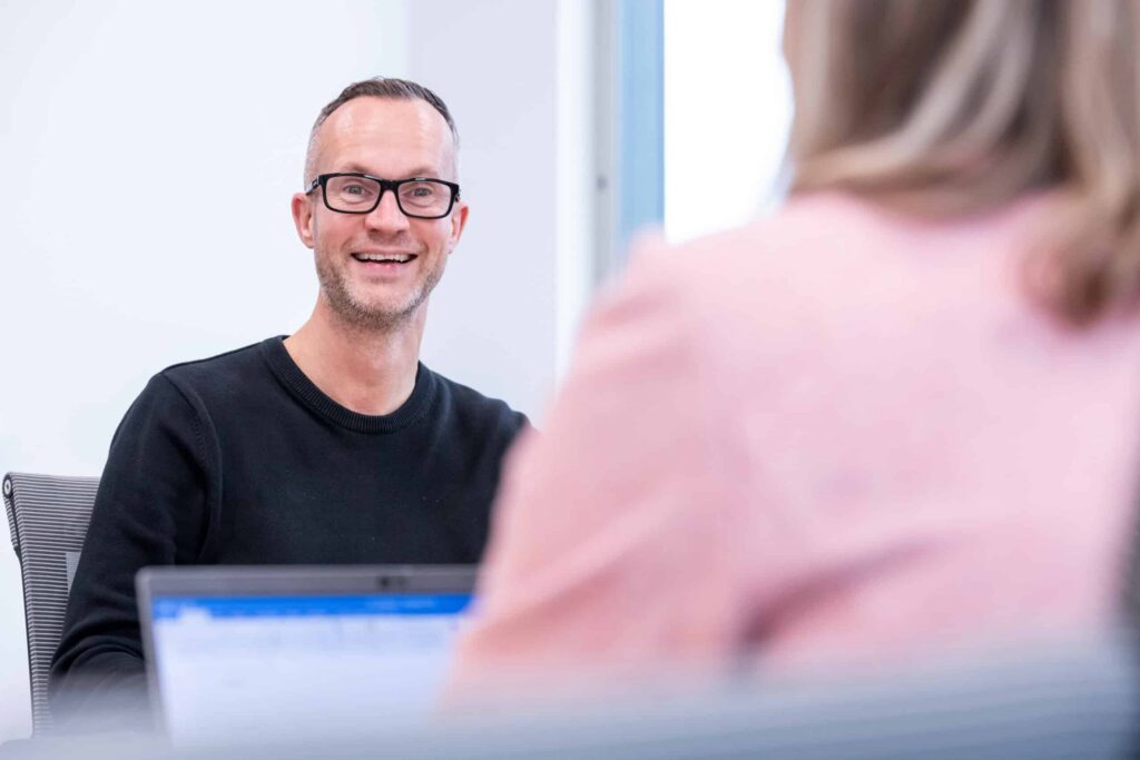 A man in glasses smiles at a colleague across the table on a laptop.
