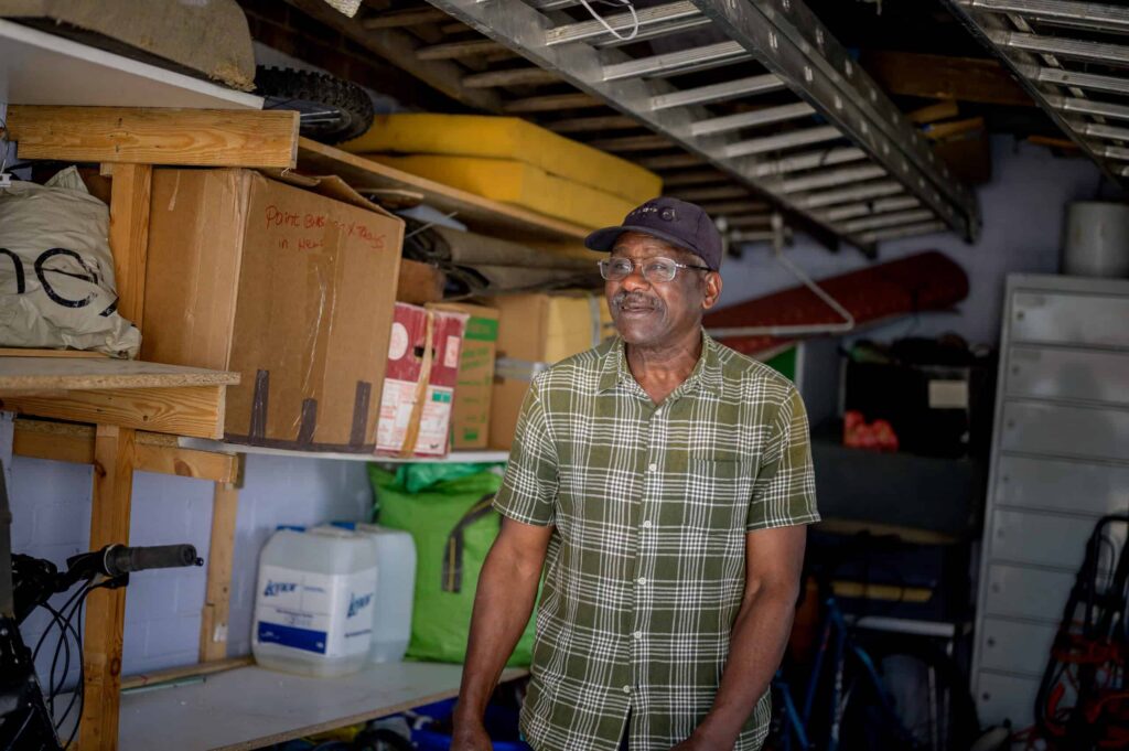 A man wearing a cap and glasses stands smiling in a shed, surrounded by boxes, a bike, washing detergent and other miscellaneous household items.