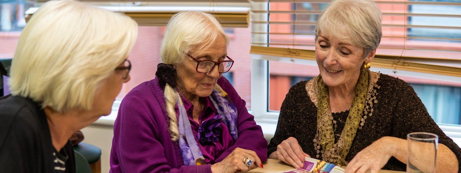 A group of three older women sitting in a room looking at a magazine in front of them.